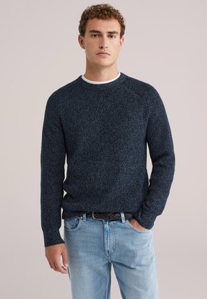 Navy gebreide trui met een gestructureerd patroon, ronde halslijn en lange mouwen, gecombineerd met lichtblauwe jeans en een zwarte riem.