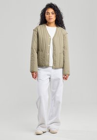 Chaqueta acolchada de color verde oliva con botones en el frente, dos bolsillos laterales y un corte holgado, llevada sobre una blusa blanca y combinada con pantalones deportivos grises.