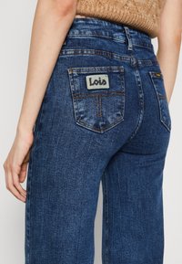 Takaa katsottuna henkilö, jolla on ylhäältä vyötärölle menevät siniset farkut, joissa on ommellut taskut, ja beige neulepusero.