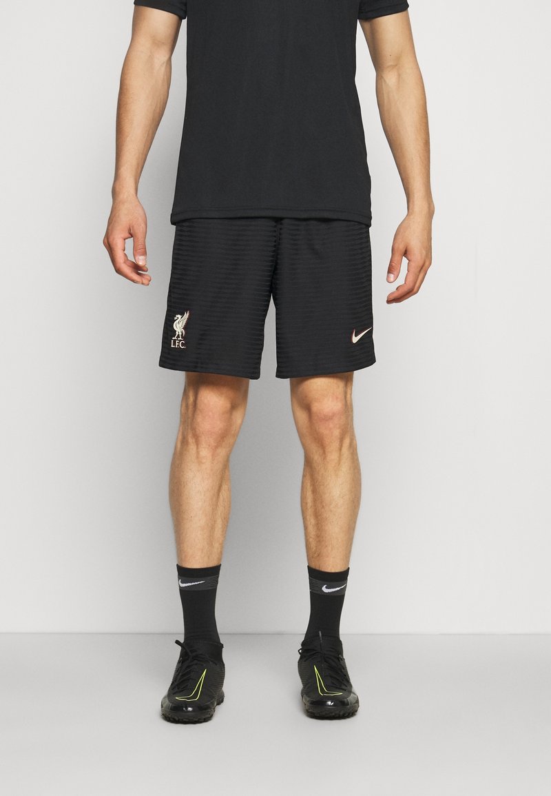 Homme portant un short noir du Liverpool FC avec logos Nike, chaussettes noires et chaussures de sport noires, debout devant un fond clair uni.