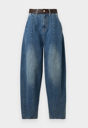 Mujer de pie con las manos en los bolsillos, lleva unos vaqueros azules de gran tamaño, una camiseta beige de manga corta con cuello en V y zapatos planos oscuros, mirando ligeramente hacia la izquierda.