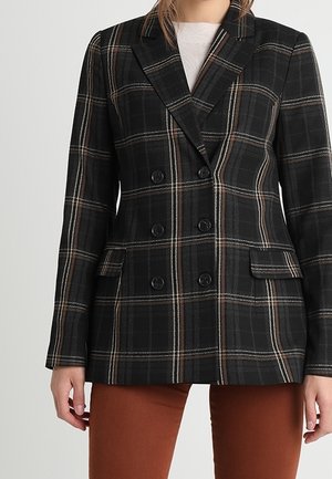 Mujer que lleva un blazer de cuadros negro de doble botonadura con líneas marrones y blancas sobre una camiseta blanca y pantalones color oxido.