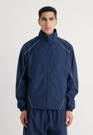Navy windbreaker jacket with a high collar, elastic cuffs, and reflective grey seams; lightweight material, front zipper closure.
