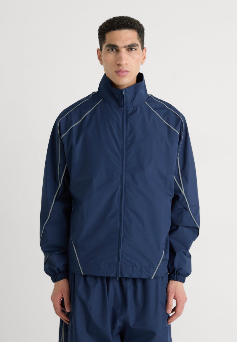 Navy windbreaker jacket with a high collar, elastic cuffs, and reflective grey seams; lightweight material, front zipper closure.