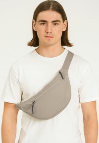 Beige Umhängetasche mit geschwungenem Design, Reißverschlusstaschen und verstellbarem Tragegurt, aus glattem, strapazierfähigem Material hergestellt.