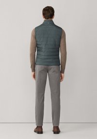 Gepolsterte dunkelgrüne Weste mit hohem Kragen und horizontaler Steppung, kombiniert mit hellgrauer Hose und dunklen Schuhen. Sauberes Design und Textur.