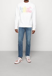 Vit sweatshirt med en flerfärgad "Endless Summer"-grafik; tillsammans med ljusblå jeans och vita sneakers med röda accenter.