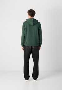 Felpa verde con cucitura posteriore e cappuccio, abbinata a pantaloni neri larghi e sneaker bianche. Texture del tessuto liscia e visibile.