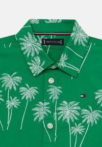 Camicia verde con bottoni, caratterizzata da stampe bianche di palme, colletto classico e una piccola patch con logo rosso, bianco e blu.
