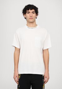 Balts kokvilnas t-krekls ar apaļu kakla izgriezumu, priekšējo kabatu un tekstu "Versace Jeans Couture" ap kaklu. Īsas piedurknes.