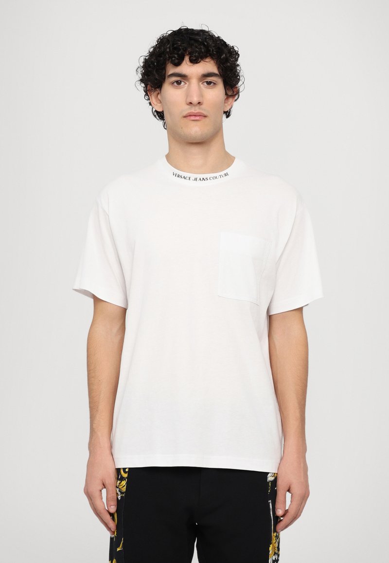 Balts kokvilnas t-krekls ar apaļu kakla izgriezumu, priekšējo kabatu un tekstu "Versace Jeans Couture" ap kaklu. Īsas piedurknes.