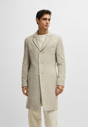 Beige Wollmischungs-Mantel mit Reverskragen, drei Knöpfen und einem subtilen strukturierten Muster, kombiniert mit einem leichten Pullover und cremefarbener Hose.