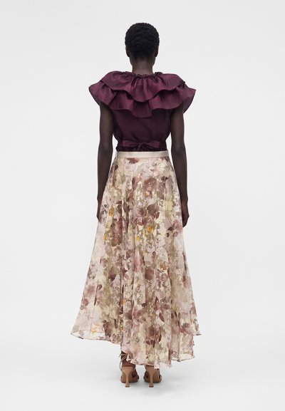 Γυναίκα που φοράει αμάνικη μπορντό μπλούζα με βολάν και μακριά, ριχτή φλοράλ φούστα με μπεζ και ροζ αποχρώσεις, με την πλάτη προς την κάμερα.