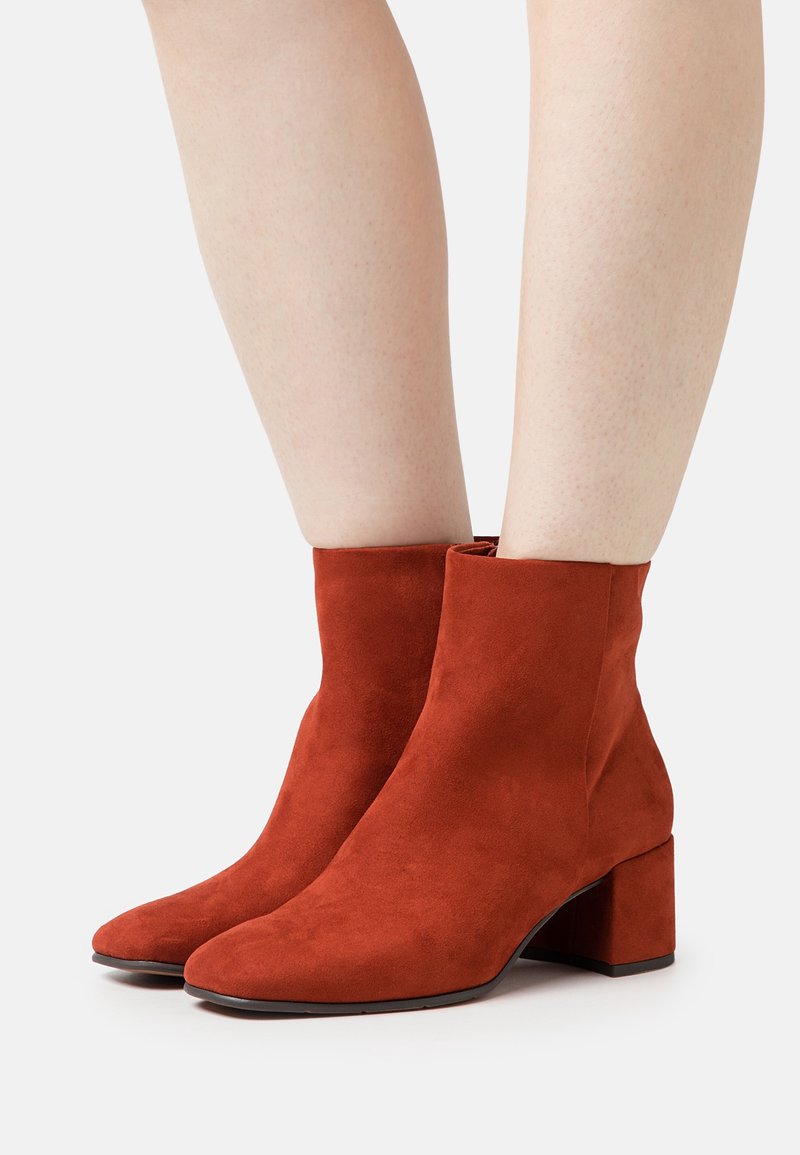 Paire de bottines en daim couleur rouille à talons carrés, portées sur des jambes nues sur fond blanc.