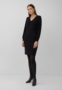 Femme debout dans une robe noire à encolure en V avec des manches longues transparentes, des collants noirs, des bottines noires et des boucles d'oreilles en or contre un fond gris.
