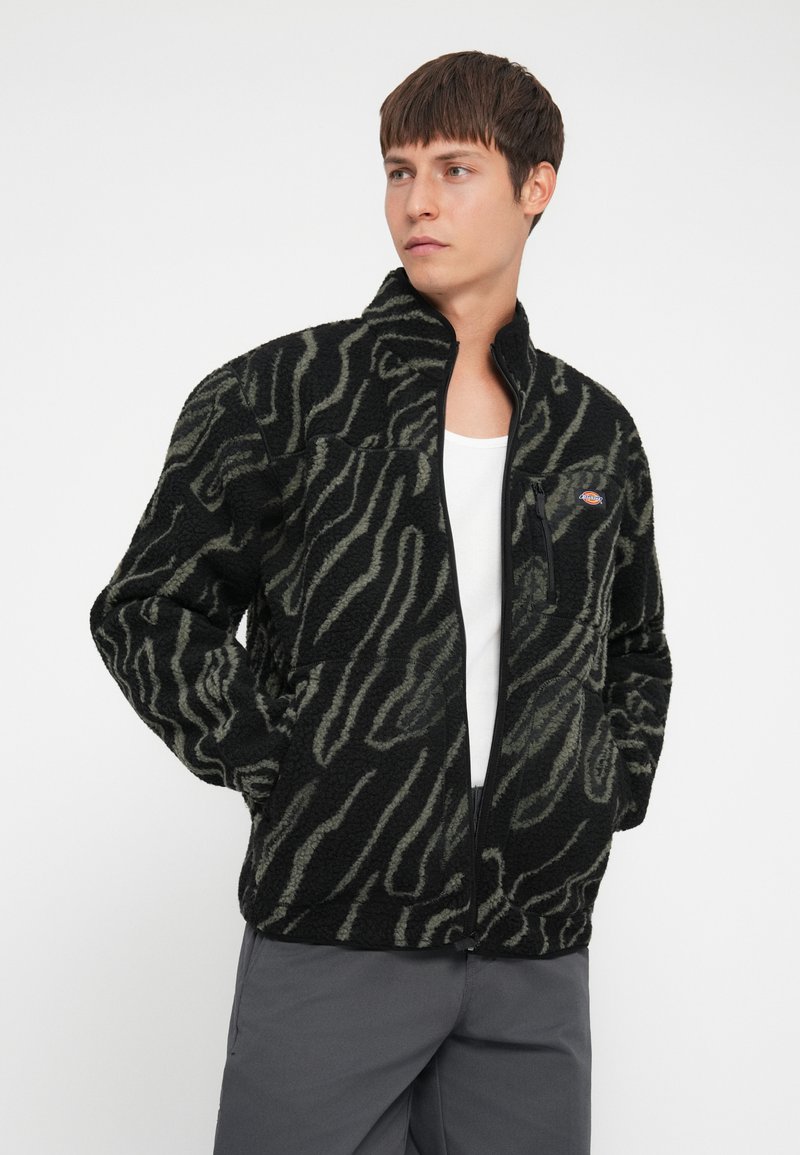 Sort fleecejakke med et grønt zebra-stribe mønster, med høj krave, frontlynlås og sidelommer.