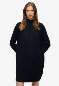 Vestido de punto azul marino con cuello alto. Presenta patrones de trenzas y dobladillos acanalados. Mangas largas con detalles texturizados.