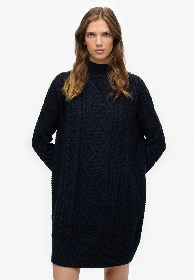 Vestido de punto azul marino con cuello alto. Presenta patrones de trenzas y dobladillos acanalados. Mangas largas con detalles texturizados.