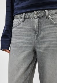 Asmuo, dėvintis šviesiai pilkas džinsines kelnes ir tamsiai mėlyną ilgomis rankovėmis marškinius su subtiliomis horizontalomis juostelėmis, ranka poilsiui prie šono.