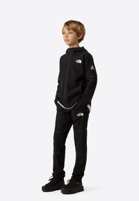 Jachetă neagră cu fermoar și pantaloni asortați, realizați dintr-un material neted și elastic. Prezintă accente cu logo și linii reflectorizante pe țesătură.