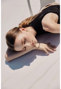 Modelo con cabello largo castaño acostada sobre una superficie clara, vistiendo una blusa negra texturada, pendientes verdes y una pulsera dorada. La luz natural resalta sus rasgos.