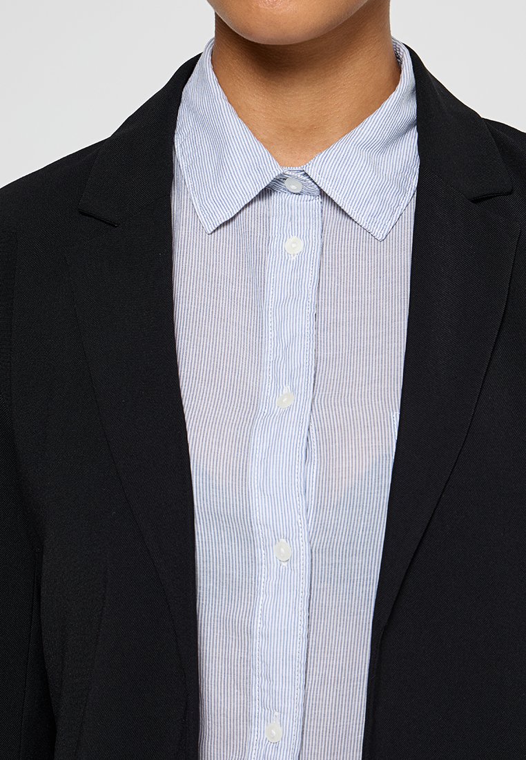 Blazer noir sur une chemise boutonné rayée bleu clair et blanche. Le col est doux, avec des boutons sur le devant et une texture de tissu lisse.