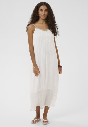 Femme aux cheveux bouclés portant une robe midi plissée blanche sans manches avec bretelles ornées et sandales tongs blanches, debout devant un fond blanc.