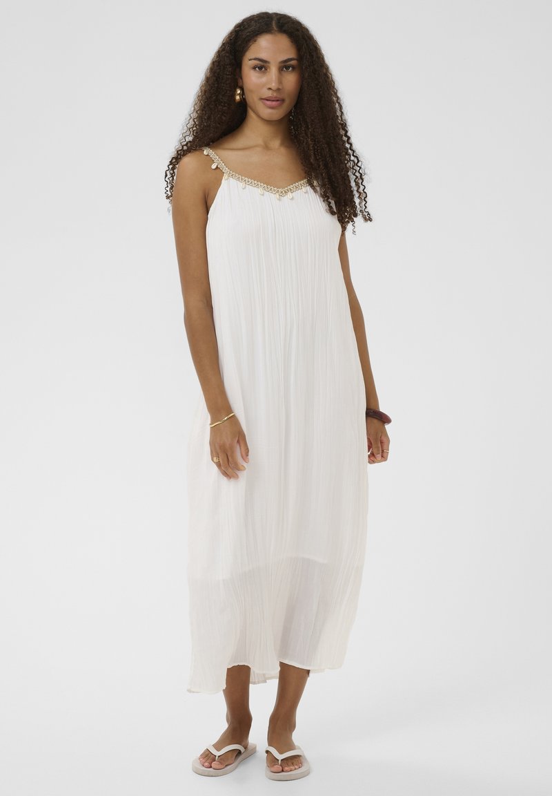 Femme aux cheveux bouclés portant une robe midi plissée blanche sans manches avec bretelles ornées et sandales tongs blanches, debout devant un fond blanc.