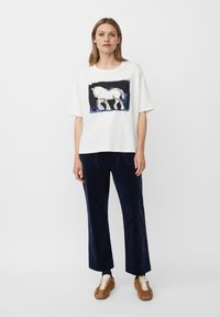 Mujer vestida con una camiseta blanca de gran tamaño con un gráfico de caballo, pantalones de pierna ancha azul marino y zapatillas marrones, de pie contra un fondo blanco liso.