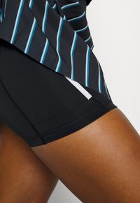 Pantalones cortos deportivos negros con tejido texturizado, que presentan un acento lateral blanco y una capa superior a rayas en tonos gris oscuro y azul.
