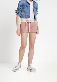 Denimtakki, valkoinen t-paita, vaaleanpunaiset shortsit, joissa on kerma-valkoinen kiristysnauha, ja valkoiset lenkkarit, yksinkertaisen vaalean taustan edessä.