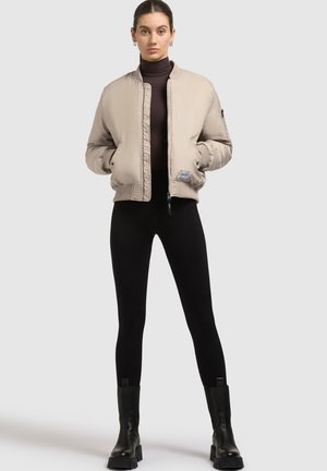 Veste bomber beige avec fermeture éclair à l'avant, poignets côtelés et détail de patch. Portée par-dessus un col roulé noir et associée avec des leggings noirs et des bottes.