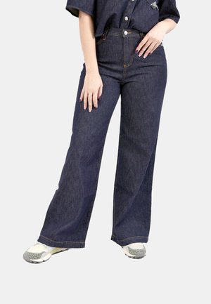 Persona che indossa jeans blu scuro a gamba larga, sneakers bianche e una camicia di denim scuro infilata, in piedi davanti a uno sfondo chiaro e uniforme.