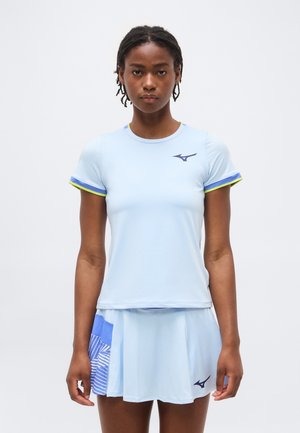 Ung kvinde iført lyseblå sportstrøje og plisseret nederdel med blåt og hvidt mønstret panel, stående foran en ensfarvet baggrund.