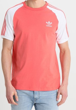 Korallinvärinen puuvillainen t-paita, jossa valkoiset raglan-hihat, ja vasemmassa rinnassa Adidas-logo sekä hihansuissa kolme raitaa.