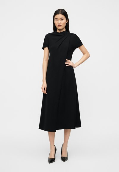 Μαύρο φόρεμα μήκους μέχρι το γόνατο με κοντά μανίκια, ψηλό λαιμό και ασύμμετρο σχέδιο τύπου wrap. Λείο ύφασμα με δομημένη εφαρμογή.