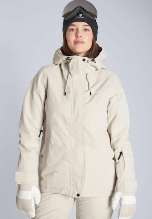 Kvinde iført beige skijakke, matchende handsker, sort hue med goggles ovenpå, står foran en ensfarvet baggrund.