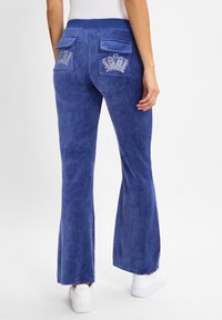 Blue velvet flared pants with a smooth texture, featuring two back pockets and embellished crown designs on each pocket.