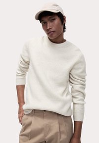 Hellbeige gestrickter Pullover mit rundem Halsausschnitt und gerippten Bündchen, kombiniert mit beigefarbenen, gefalteten Hosen und einer beigefarbenen Mütze.