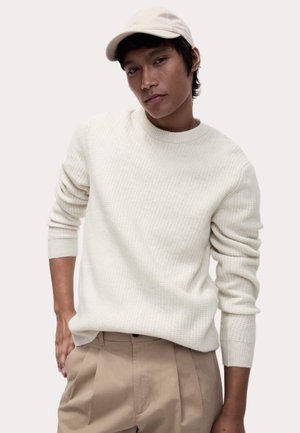 Hellbeige gestrickter Pullover mit rundem Halsausschnitt und gerippten Bündchen, kombiniert mit beigefarbenen, gefalteten Hosen und einer beigefarbenen Mütze.