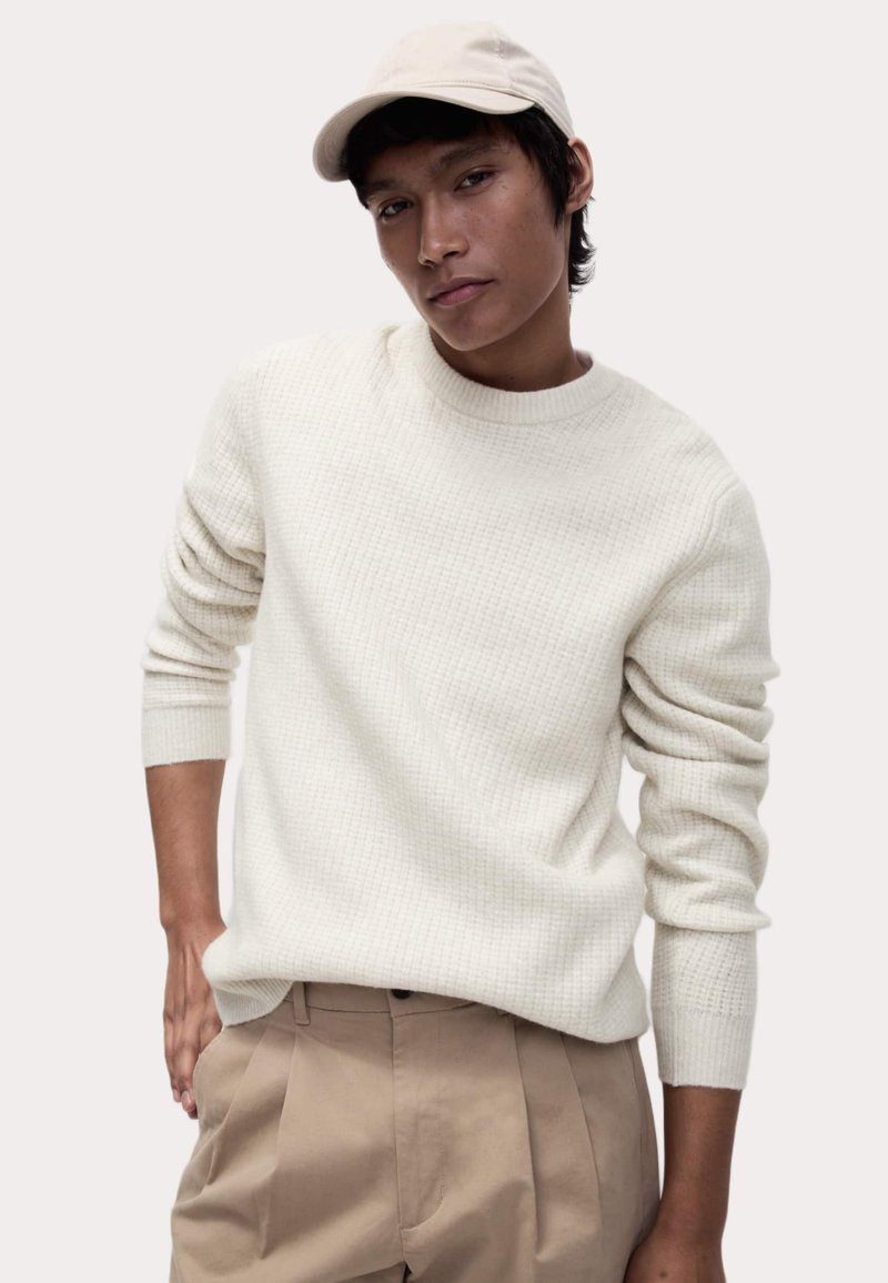 Hellbeige gestrickter Pullover mit rundem Halsausschnitt und gerippten Bündchen, kombiniert mit beigefarbenen, gefalteten Hosen und einer beigefarbenen Mütze.