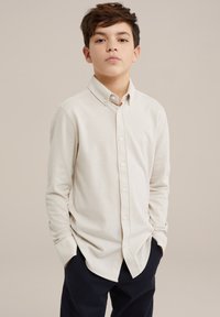 Camisa de manga larga beige claro con cuello abotonado, botones en la parte delantera y una textura suave, combinada con pantalones oscuros. Diseño simple.