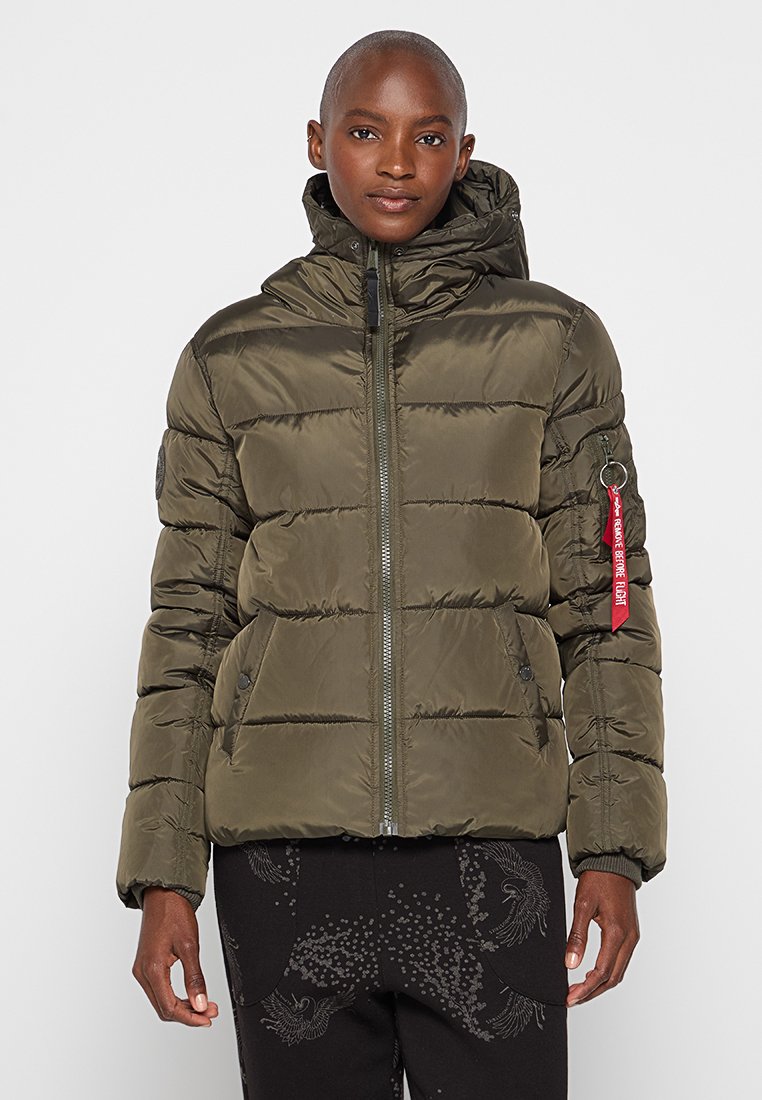 Chaqueta acolchada verde oliva con cierre frontal, capucha y bolsillos con cremallera. Presenta costuras horizontales y un acento de etiqueta roja en la cremallera.