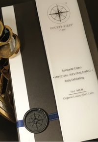 Lichaamspeelingcrème in een witte tube, met een zwart logo, verzegeld met een marineblauwe lint en een zwart waszegel, geplaatst op donkere verpakking.