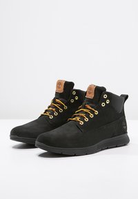 Botas de tobillo de ante negro con suela de goma, cordones amarillos y ojetes dorados. Presenta una etiqueta de cuero marrón y un panel texturizado en el lateral.