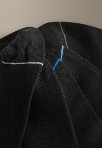 Quatre paires de chaussettes noires disposées en chevauchement, chacune avec une seule ligne de couture colorée en blanc, bleu ou gris près de l'orteil.