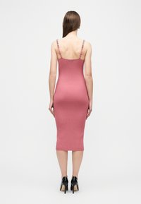 Γυναίκα με μακριά μαλλιά που φοράει εφαρμοστό, ριμπ πλεκτό, ροζ μίντι φόρεμα με λεπτές τιράντες και μαύρα ψηλοτάκουνα παπούτσια, στέκεται με την πλάτη προς τα εμπρός.