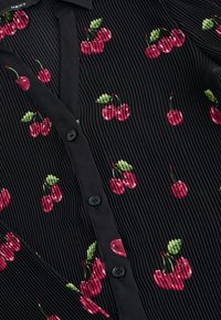 Chemise noire en tissu côtelé à manches courtes, ornée d'une broderie de cerises rouges avec des feuilles vertes, détaillée avec des boutons noirs sur le devant.