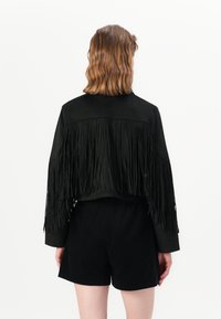 Čierna semišová skrátená bunda s dlhými fringingmi na rukávoch, spárovaná s čiernymi šortkami. Dizajn je minimalistický bez viditeľných zapínaní.