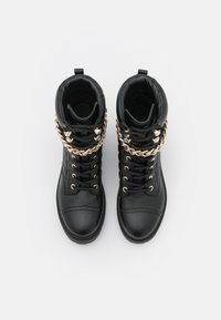 Botas de tobillo de cuero negro con acentos de cadena metálica dorada, punta redonda, cuello acolchado y parte delantera con cordones. Superficie texturizada con detalles de costura.